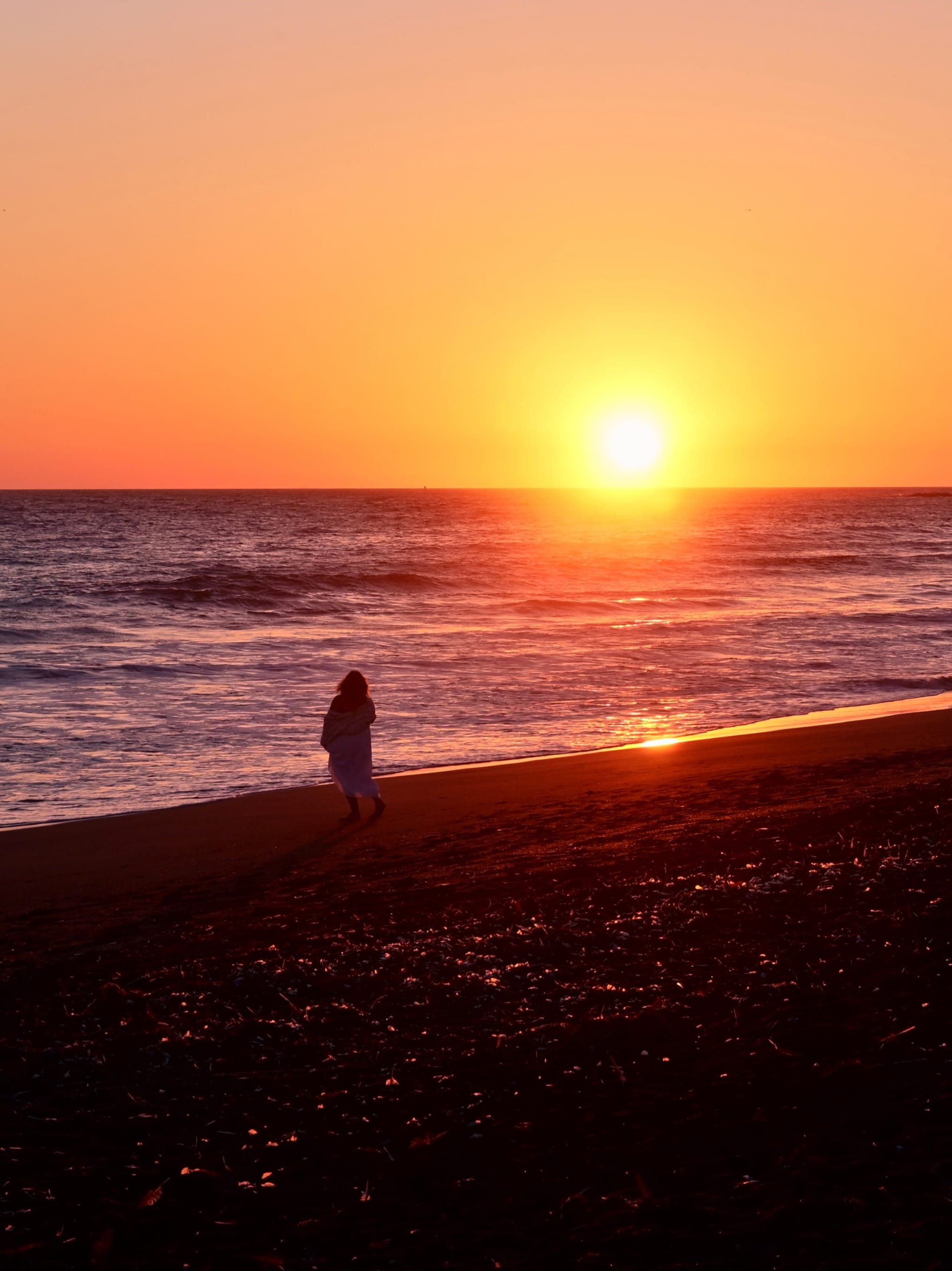 A person walking along a beach at sunset, with the sun low on the horizon and the sky filled with warm orange hues
