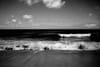 A black and white seascape with waves crashing onto the shore under a cloudy sky
