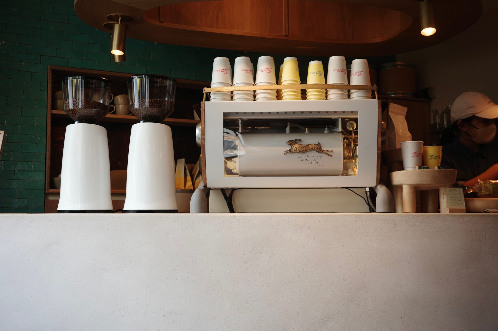 A coffee shop counter with a white espresso machine, cups stacked on top, and two grinders on the left