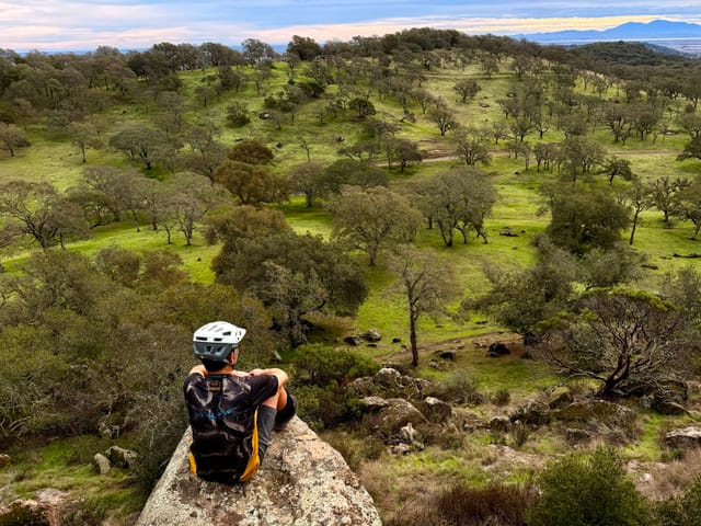 Cyclist with a backpack and helmet sits on a large rock overlooking rolling green hills dotted with trees under a cloudy sky
