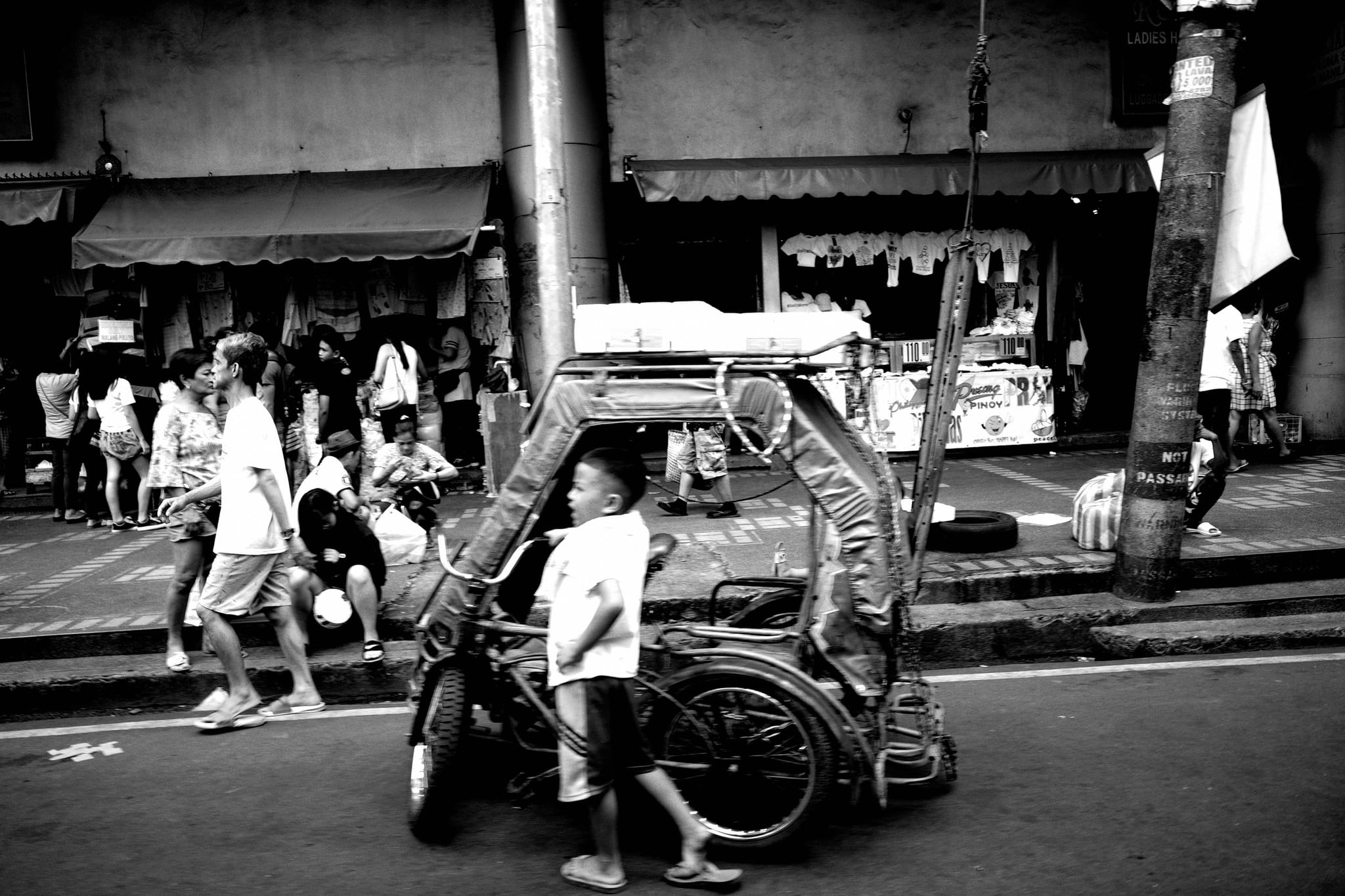 A black and white street scene with a child standing next to a tricycle. People walk by, and shops are visible in the background