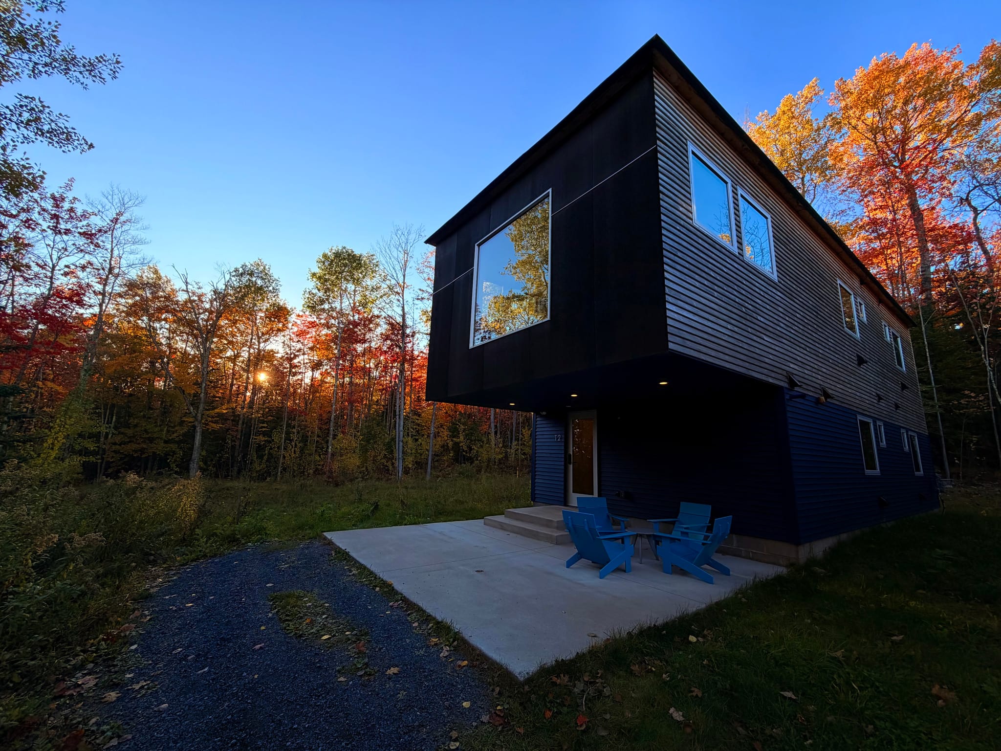 Modern dark-clad cabin with a cantilevered upper floor and large window, a small patio with blue Adirondack chairs, set in a wooded area with autumn foliage at sunset