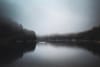 A tranquil lake bordered by dense, misty forests under an overcast sky, creating a serene and mysterious atmosphere with reflections on the water