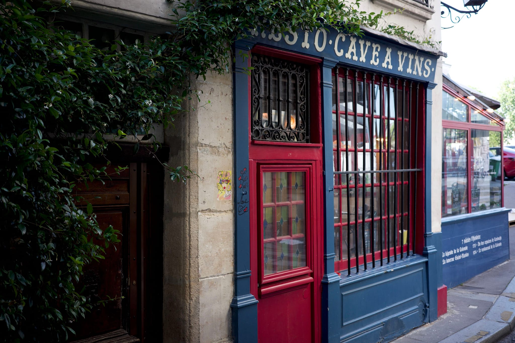 A quaint storefront with a red door and window frames, featuring the words BISTRO CAVE A VINS above. The building has a classic, rustic appearance with greenery on the side