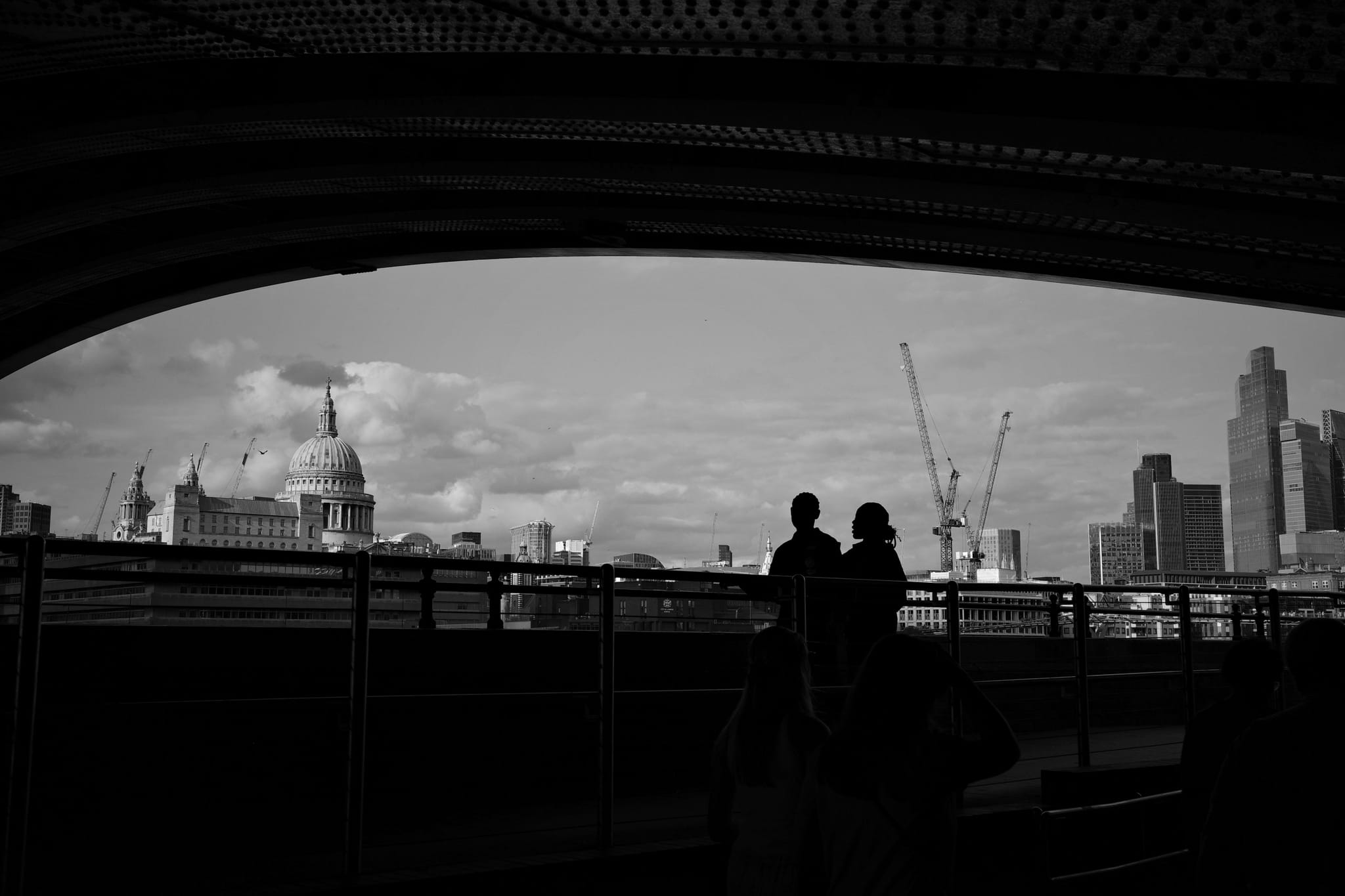 A black and white silhouette of two people standing under a bridge, with a cityscape in the background featuring cranes and a prominent dome