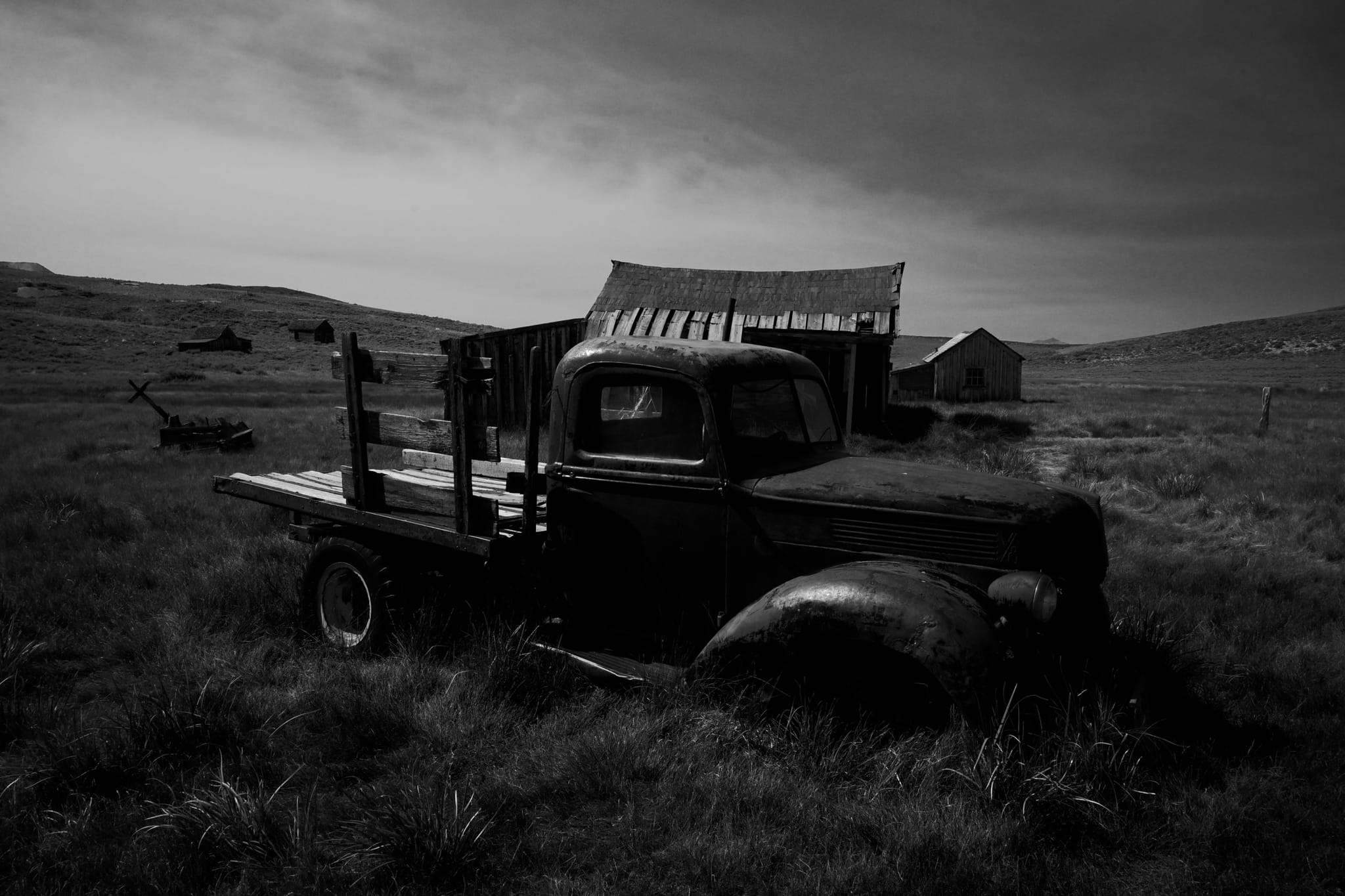A black and white scene featuring an old, abandoned truck in a grassy field with a rustic barn and distant hills under a cloudy sky
