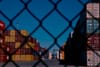 Stacks of colorful shipping containers are visible through a chain-link fence under a clear blue sky