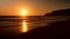 A serene sunset over the ocean with gentle waves lapping against a sandy beach and a silhouette of a distant hill