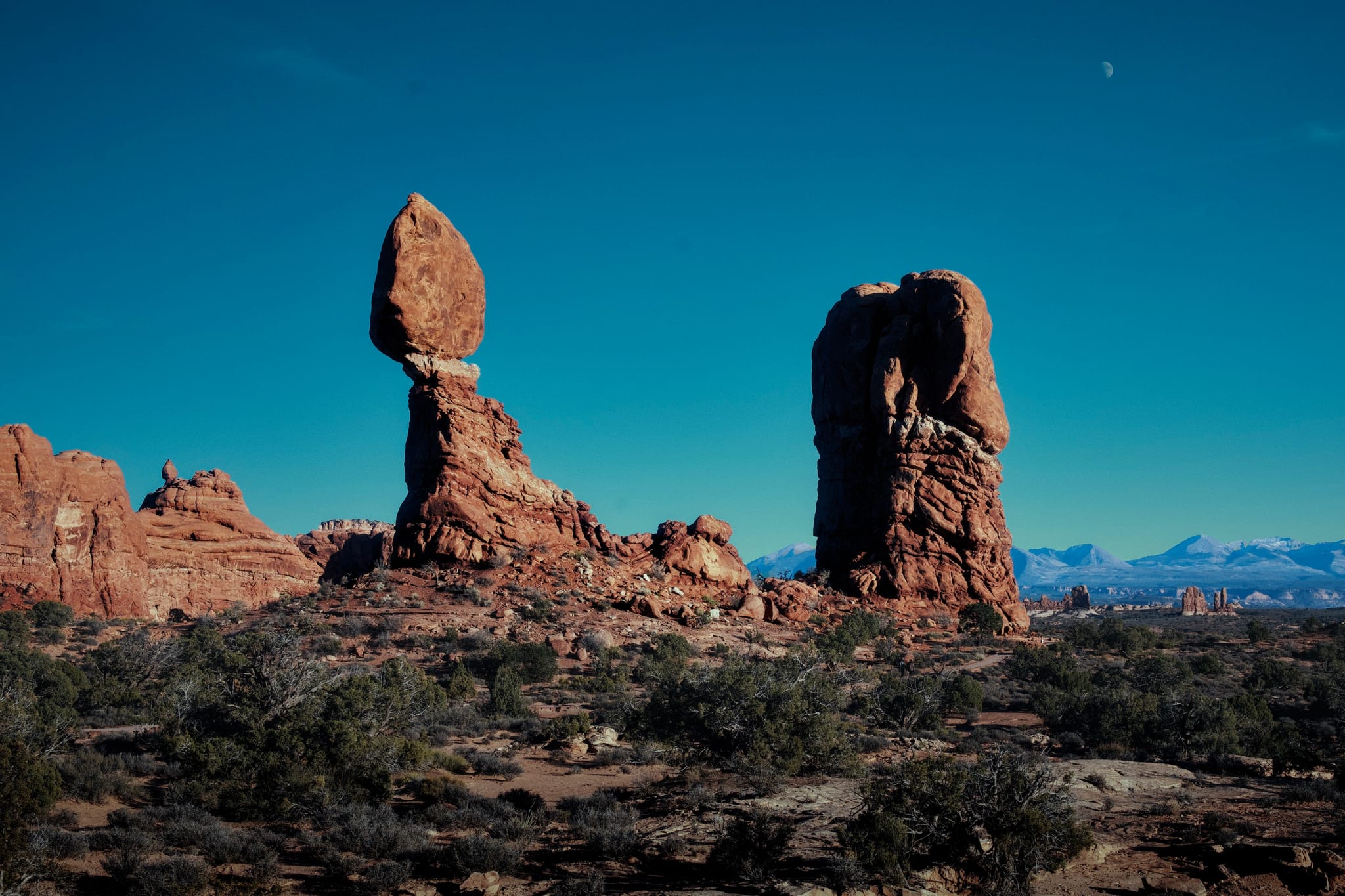 Balanced rock formation and sandstone spire in a desert landscape with distant snowcapped mountains under a clear blue sky