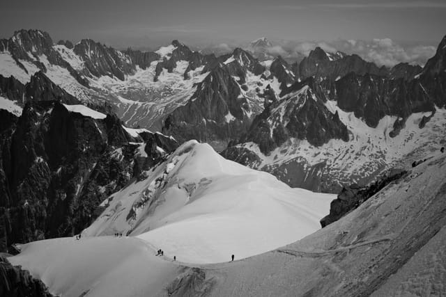 A black and white photograph of a snowy mountain range with rugged peaks and a few climbers traversing a snow-covered ridge
