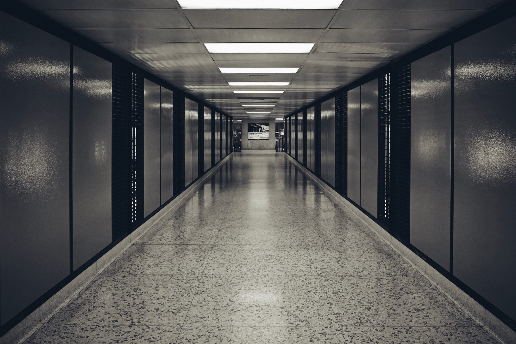 A long, empty corridor with a polished floor, flanked by dark walls and illuminated by overhead fluorescent lights
