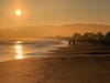 A serene beach scene at sunset with the sun low in the sky, casting a warm glow over the water and sand. Silhouettes of people are visible walking along the shoreline, with distant hills in the background