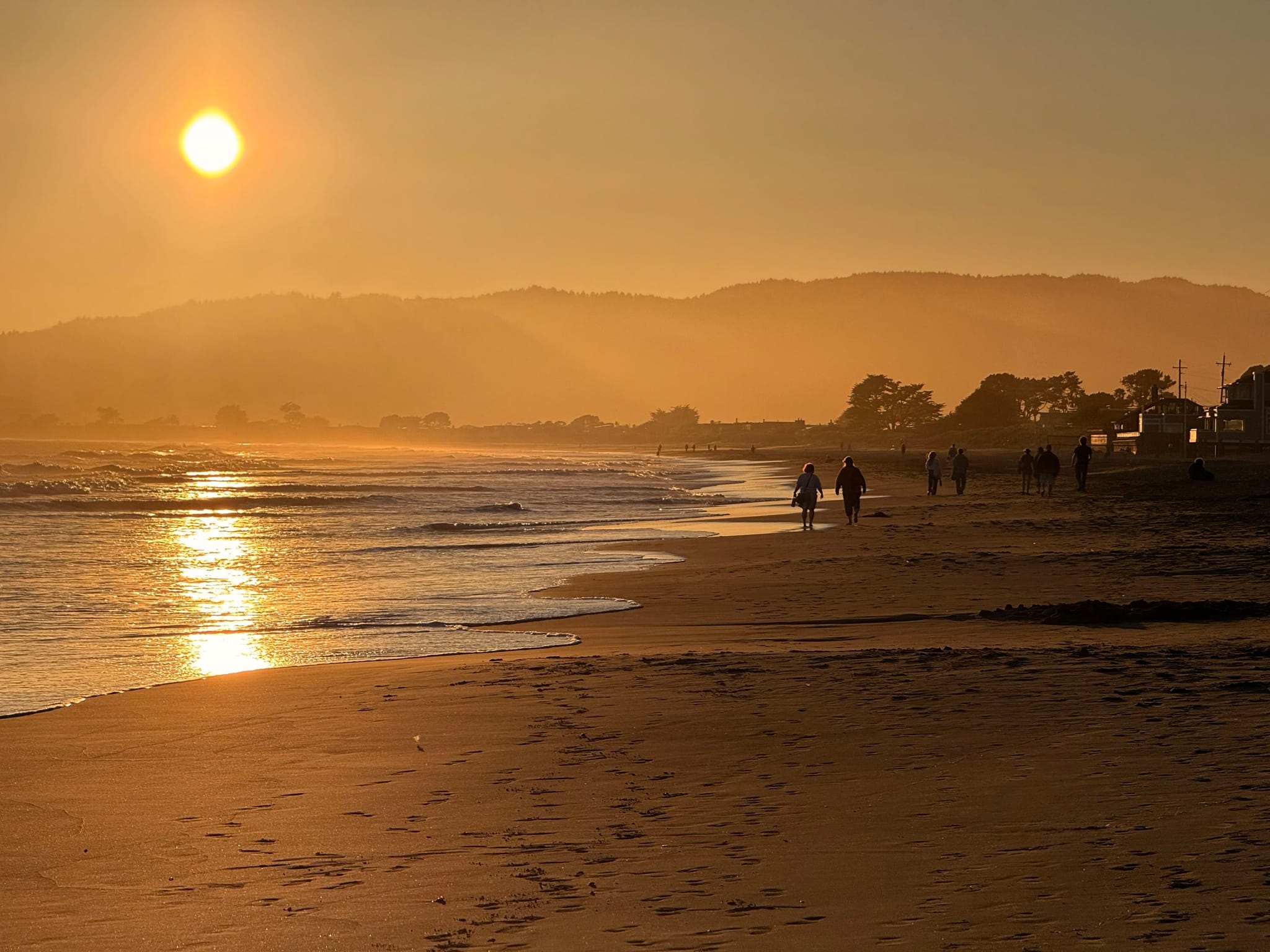 A serene beach scene at sunset with the sun low in the sky, casting a warm glow over the water and sand. Silhouettes of people are visible walking along the shoreline, with distant hills in the background