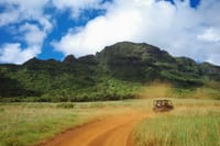 A rugged vehicle drives along a dirt road through a grassy landscape, with a lush green mountain and a partly cloudy sky in the background