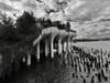 Black-and-white view of Little Island on the Hudson River, its tulip-shaped concrete columns standing above the water amid old wooden pilings under a dramatic cloudy sky