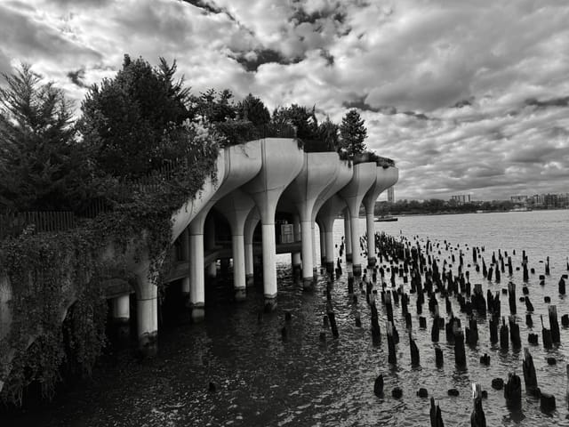Black-and-white view of Little Island on the Hudson River, its tulip-shaped concrete columns standing above the water amid old wooden pilings under a dramatic cloudy sky