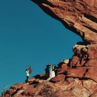 Wedding couple and photo crew on red sandstone ledge beneath a large natural arch against clear blue sky