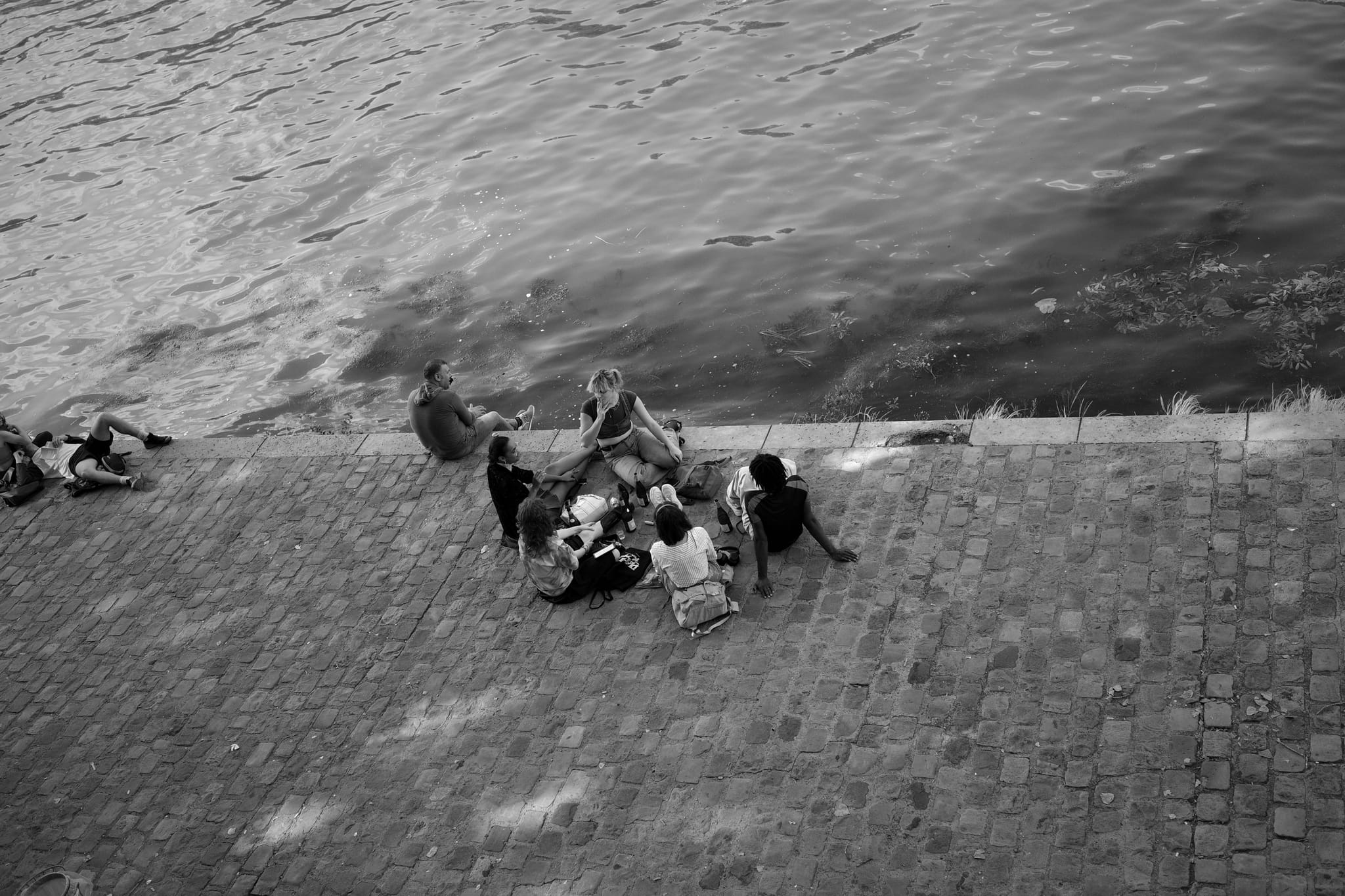 A black and white scene of people sitting on a cobblestone path by the water, engaged in conversation and relaxation