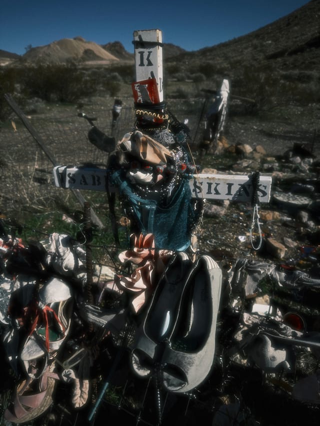 Roadside memorial cross surrounded by flowers, candles, and ski gear in a desert roadside setting
