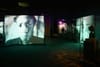 A dimly lit exhibition space with large screens displaying black and white portraits, creating an immersive atmosphere
