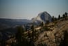 A mountainous landscape with a prominent granite dome in the background, surrounded by trees and rocky terrain under a clear sky