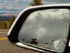 Approaching pickup truck reflected in a car’s side-view mirror on a scenic highway, with dramatic clouds and distant hills