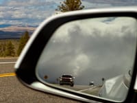 Approaching pickup truck reflected in a car’s side-view mirror on a scenic highway, with dramatic clouds and distant hills