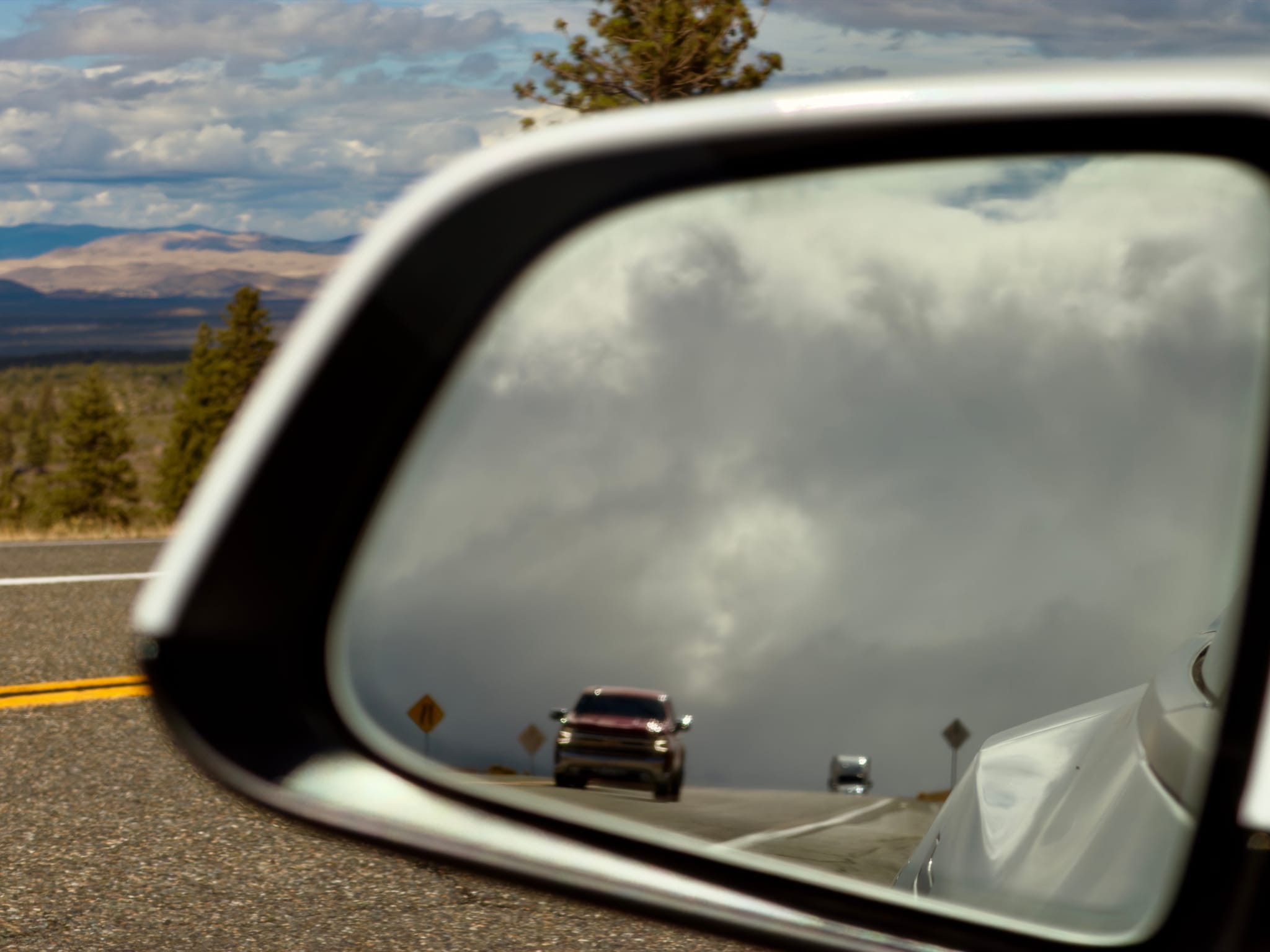 Approaching pickup truck reflected in a car’s side-view mirror on a scenic highway, with dramatic clouds and distant hills