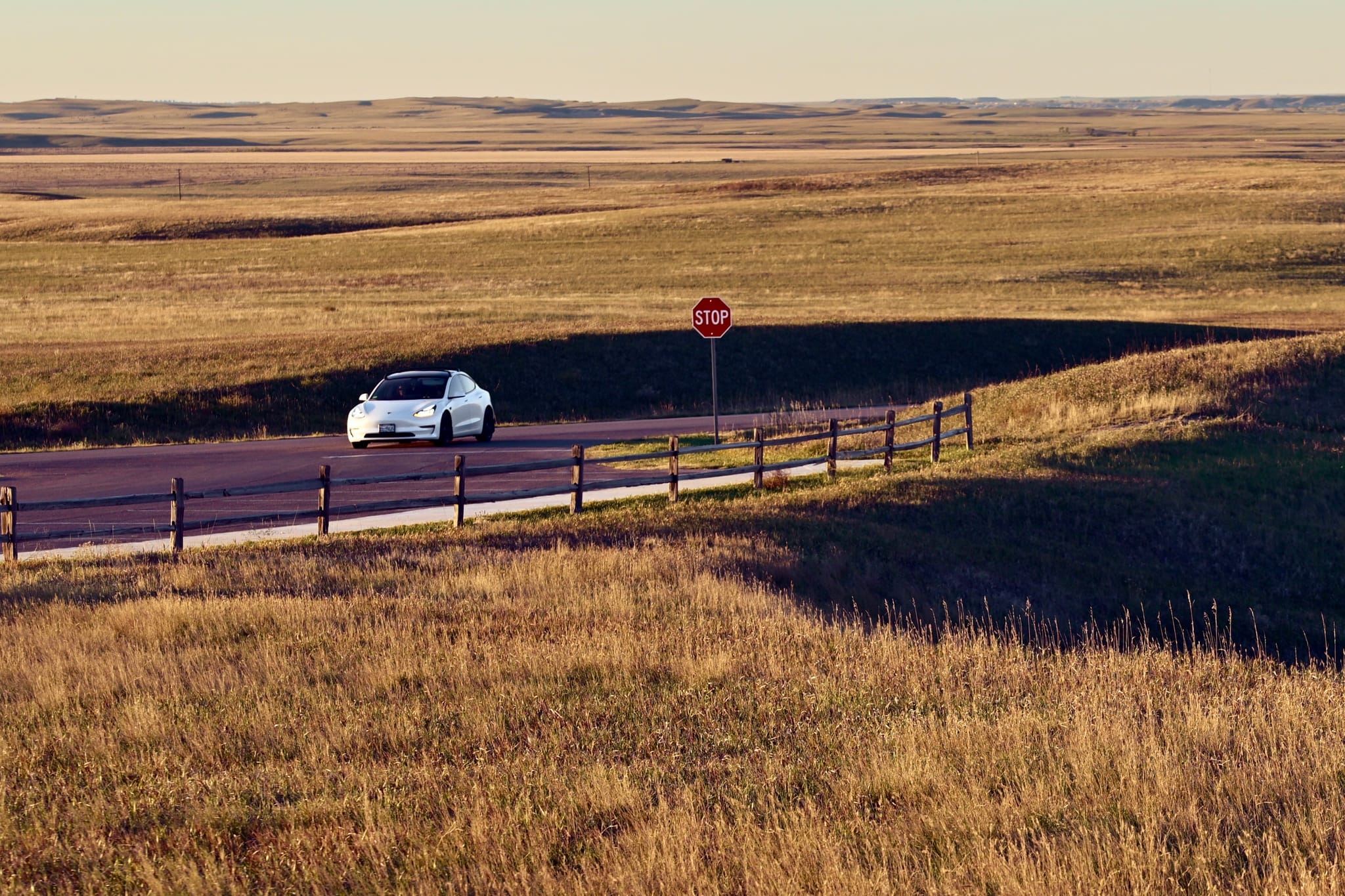 A white car on a lonely road cutting through golden grassland, passing a stop sign and wooden fence in late-afternoon light