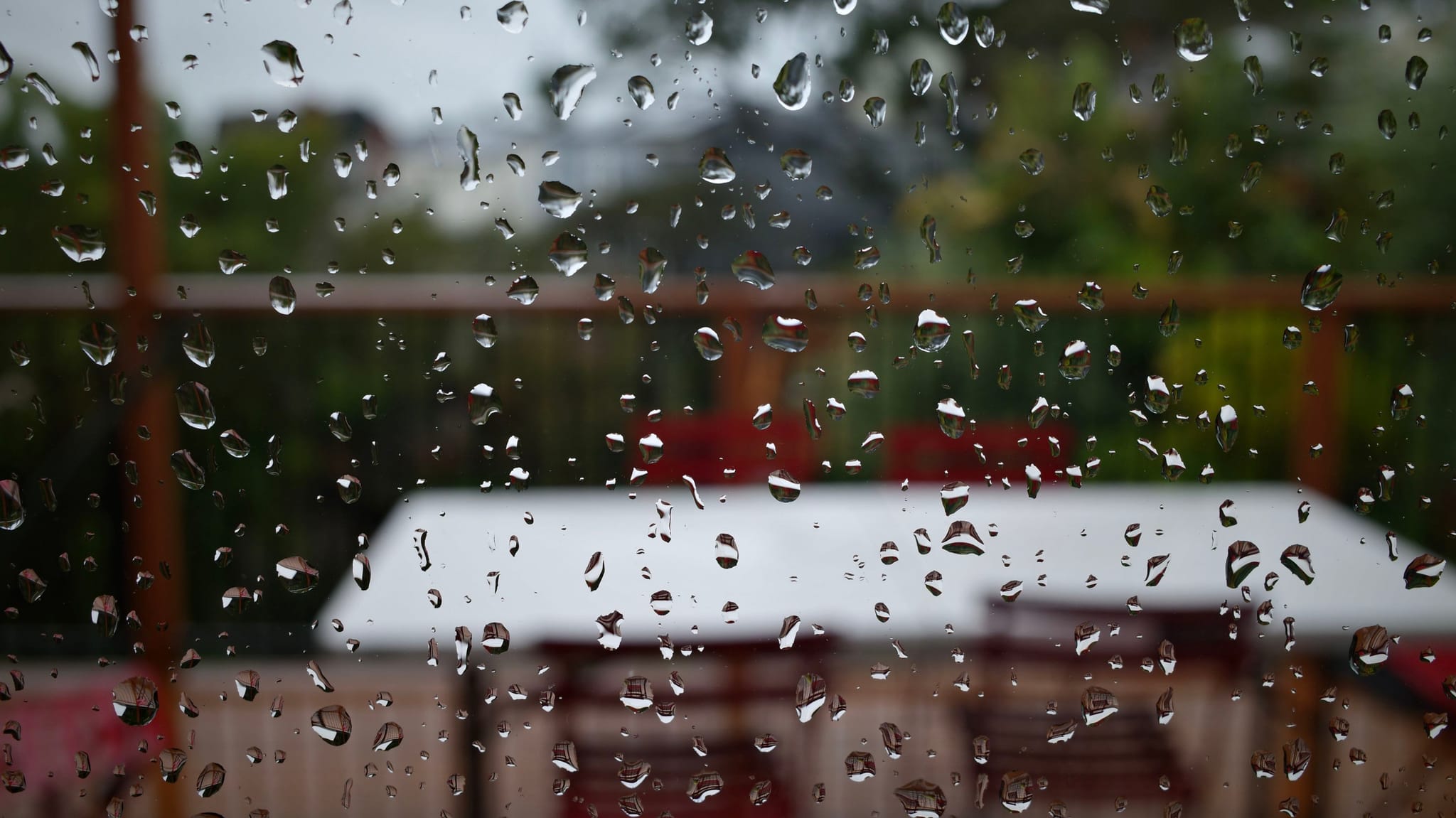 Raindrops on a window with a blurred view of outdoor furniture and greenery in the background