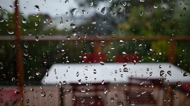 Raindrops on a window with a blurred view of outdoor furniture and greenery in the background