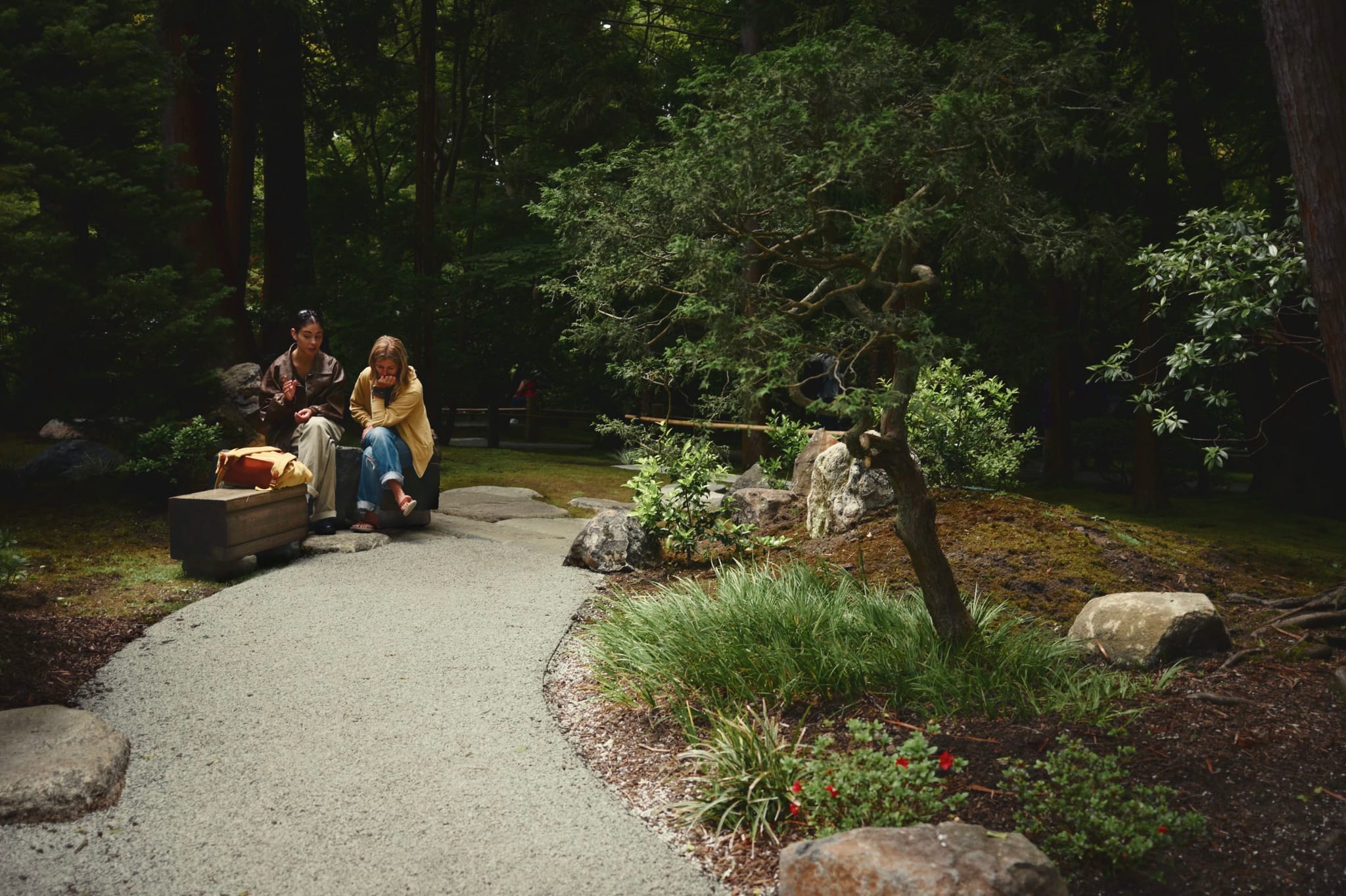 A serene garden scene with a winding gravel path, surrounded by lush greenery and rocks. Three people are sitting on a bench to the left, engaged in conversation