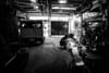 A black and white scene inside a workshop with a person working on a project, surrounded by tools and equipment. A vehicle is parked on the left, and light filters in from the open entrance