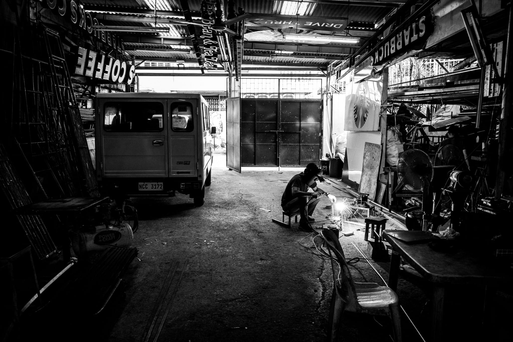A black and white scene inside a workshop with a person working on a project, surrounded by tools and equipment. A vehicle is parked on the left, and light filters in from the open entrance