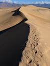 Sand dune crest with a line of footprints along the ridge, casting a long shadow across the slope with distant mountains on the horizon