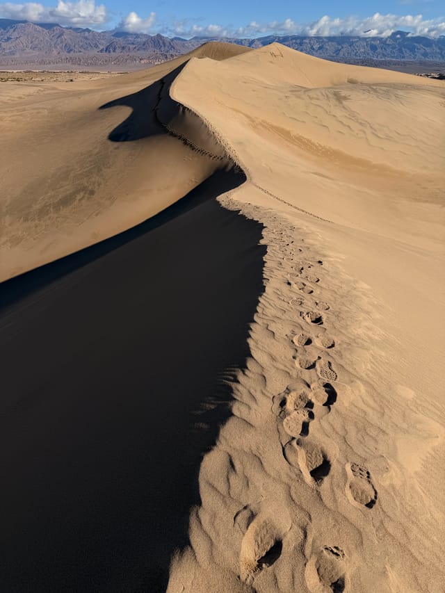Sand dune crest with a line of footprints along the ridge, casting a long shadow across the slope with distant mountains on the horizon