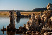A serene lake with unique rock formations rising from the water, surrounded by a flat landscape and distant hills under a clear blue sky