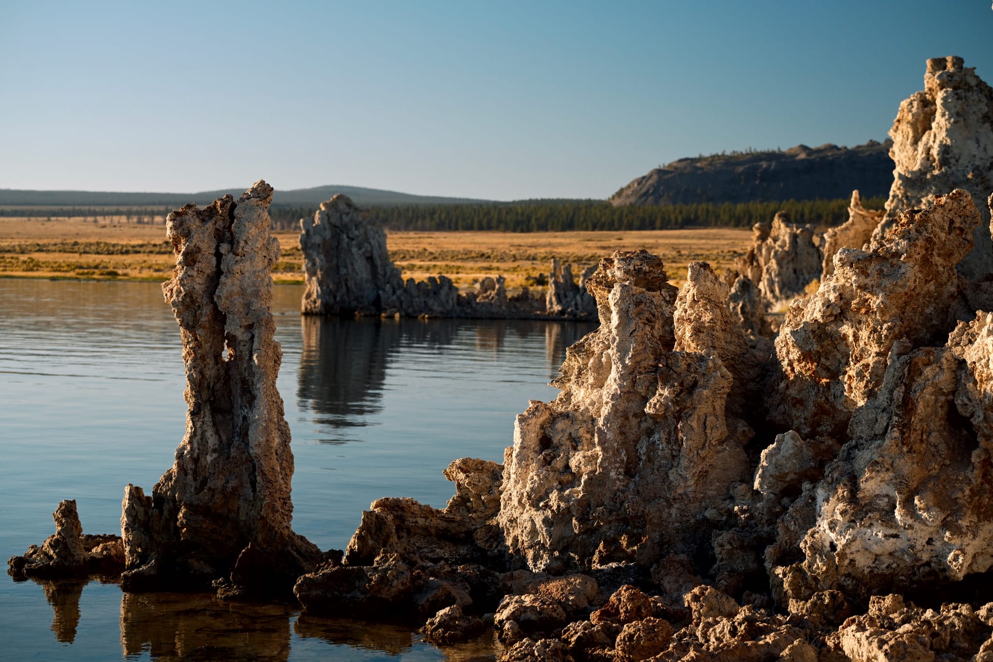 A serene lake with unique rock formations rising from the water, surrounded by a flat landscape and distant hills under a clear blue sky