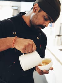 A person wearing a beanie and dark clothing is skillfully pouring milk from a jug into a cup, likely making latte art