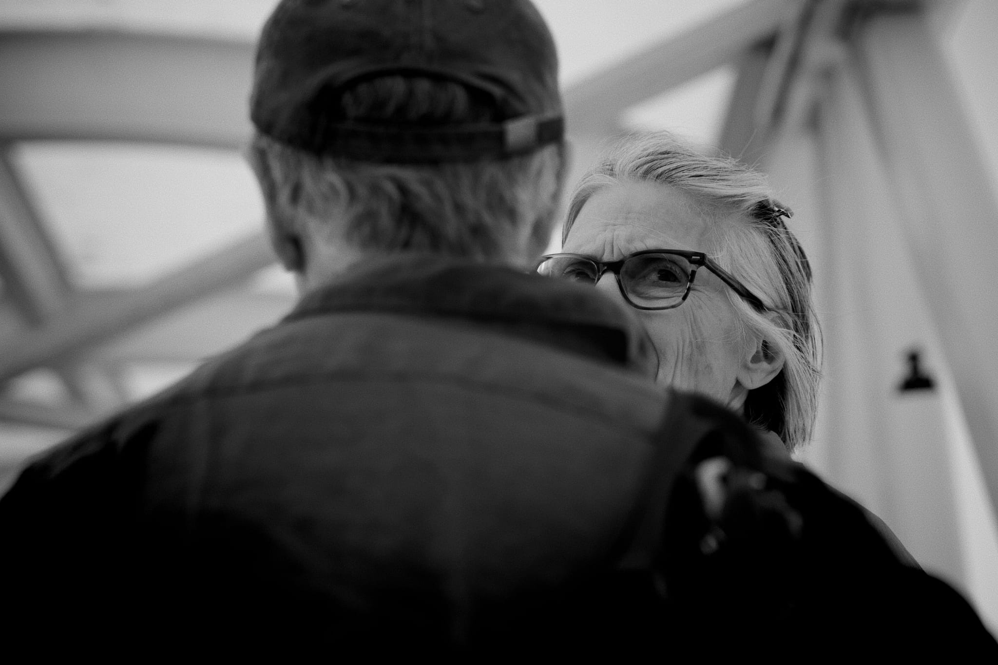 A black and white photo of two people facing each other, one wearing glasses and the other a cap, with a blurred background