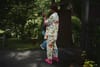 A person wearing colorful floral pajamas and pink shoes stands outdoors, holding a camera. The background features greenery and a wooden structure