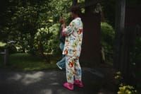 A person wearing colorful floral pajamas and pink shoes stands outdoors, holding a camera. The background features greenery and a wooden structure