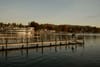 Wooden pier extending into a calm lake at sunset, with a moored ferry and lakeside buildings backed by autumn-colored hills