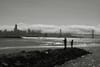A black and white scene featuring two people standing on a rocky shoreline with a city skyline and a bridge in the background, under a cloudy sky