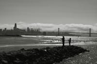 A black and white scene featuring two people standing on a rocky shoreline with a city skyline and a bridge in the background, under a cloudy sky