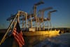 A group of large industrial cranes on a dock, with an American flag in the foreground and water reflecting the golden light of sunset