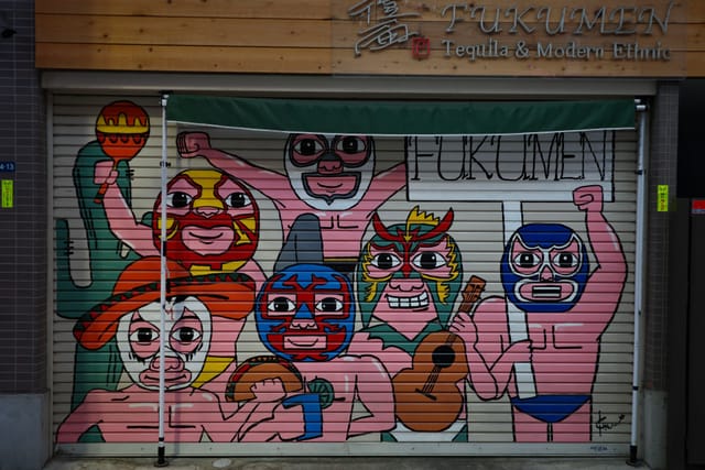 A colorful mural on a metal shutter featuring cartoonish characters wearing luchador masks, each with different expressions and poses, with the word FUKUMEN above them