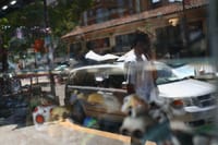 A person walking past a parked car on a sunny street, with reflections of various objects in a window