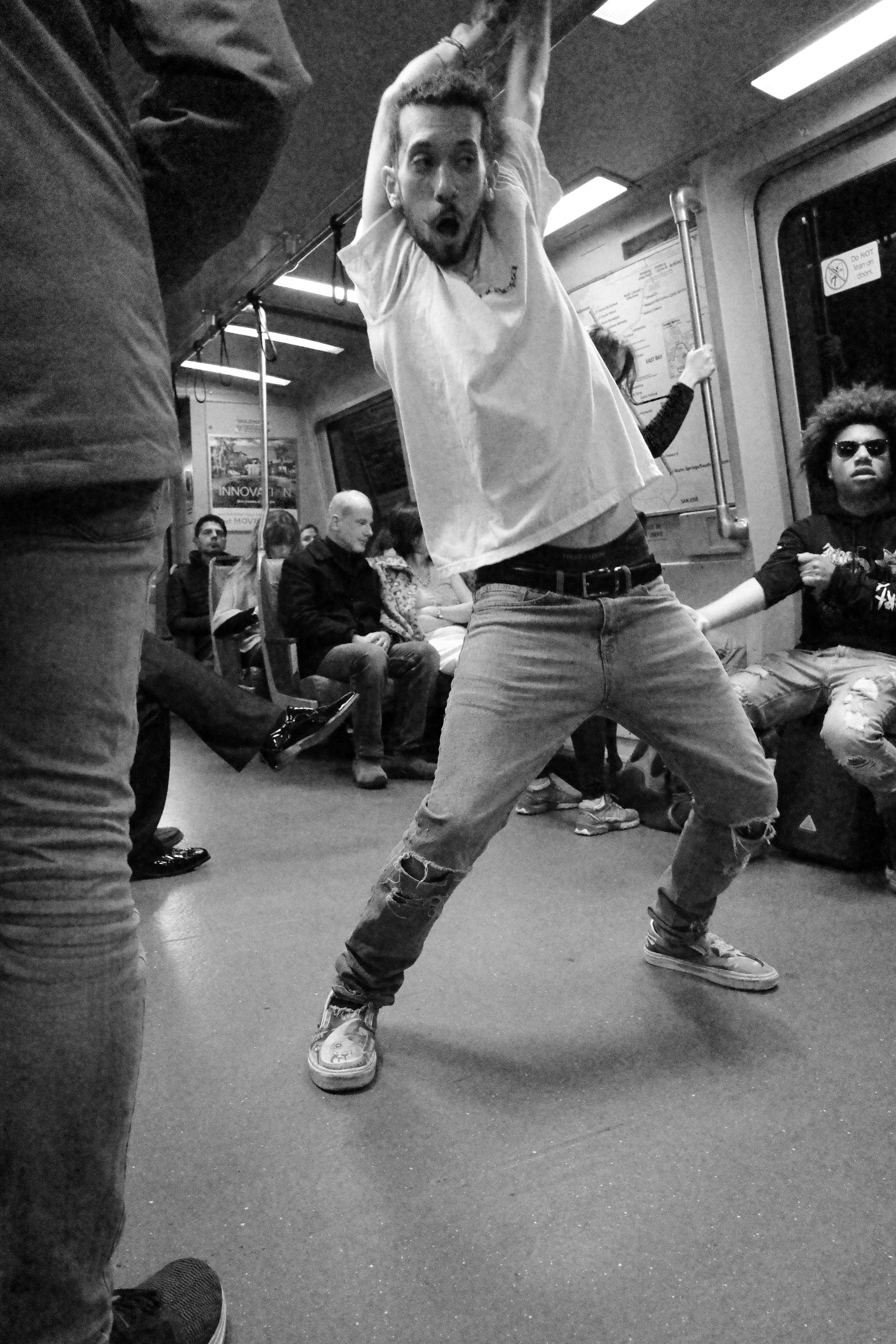 A person striking a dynamic pose in a subway car, surrounded by seated passengers. The scene is captured in black and white