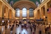 A bustling interior of a grand train station with high arched windows, a large American flag, and numerous people moving about